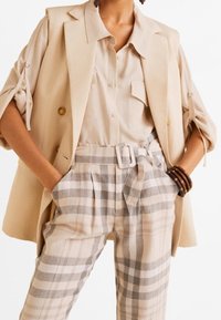 Beige dvouřadý blazer s ohrnutými rukávy, zkombinovaný s světle béžovou košilí a kostkovanými kalhotami s vysokým pasem a páskem.
