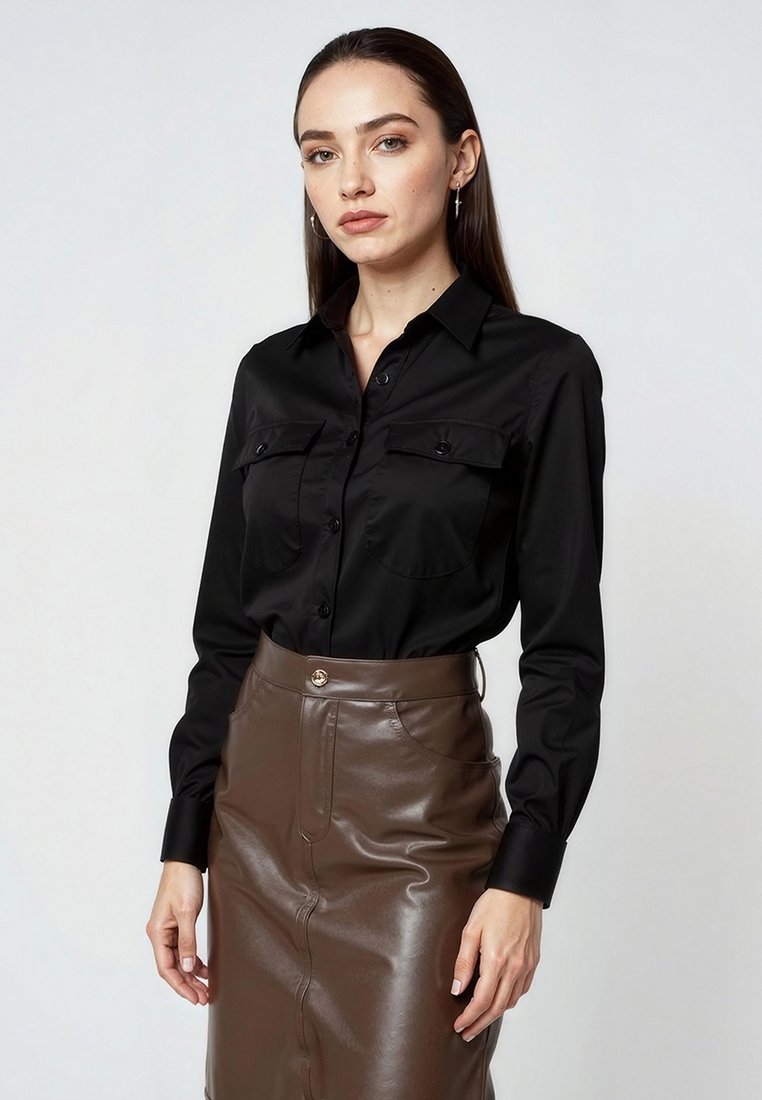 Chemise noire à manches longues avec un devant à boutons, deux poches poitrine et col pointu. Associée avec une jupe en cuir brun ajustée.