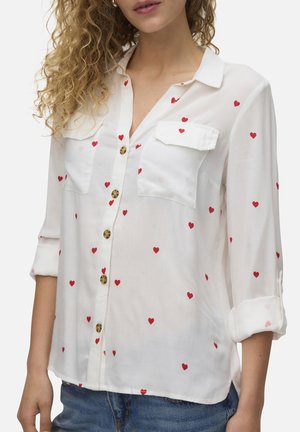 Femme portant une chemise blanche à manches longues boutonnée avec des motifs de cœurs rouges et des manches retroussées, associée à un jean bleu.