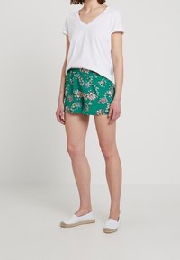 T-shirt bianco con scollatura a V abbinato a pantaloni corti verde con stampa floreale, caratterizzati da una vestibilità comoda e una cintura elastica. Scarpe bianche slip-on con dettagli in paglia.