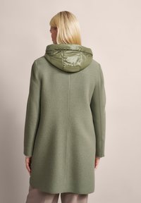 Grüner Mantel aus einem strukturierten, wollähnlichen Außenmaterial und einer glänzenden, gesteppten Kapuze. Lange Ärmel und ein leicht geschwungener Saum hinten.