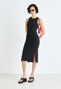 Μαύρο αμάνικο φόρεμα με πλαϊνή σχισμή, φτιαγμένο από ριμπ ύφασμα. Το μοντέλο φορά μαύρες πλατφόρμες και κρατά μια κόκκινη τσάντα.