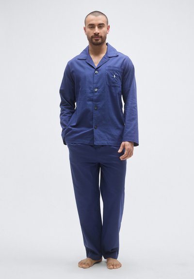 Σετ πιτζάμες από βαμβάκι σε ναυτικό μπλε χρώμα με μακριά μανίκια, κούμπωμα με κουμπιά και τσέπη στο στήθος. Το παντελόνι έχει άνετη εφαρμογή· το υλικό φαίνεται λείο και απαλό.