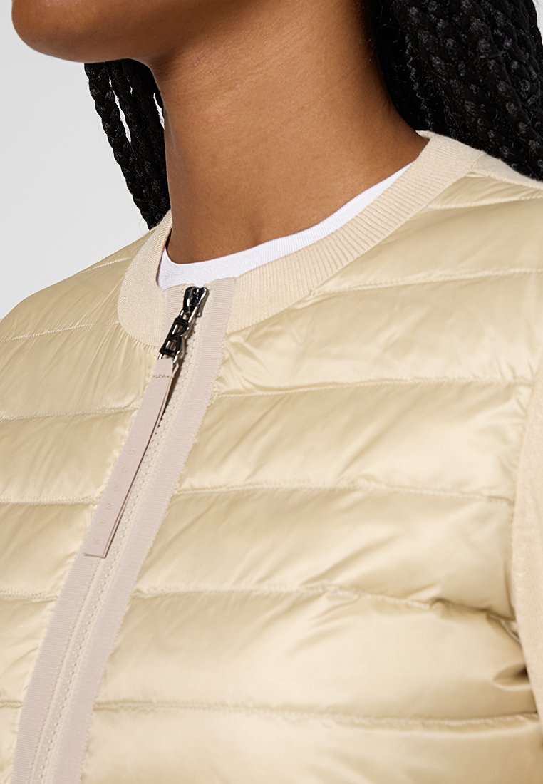 Beige Pufferjacke mit gestepptem Design, ausgestattet mit einem Stehkragen und einem Reißverschluss. Der Stoff ist glatt und wird durch Strickbesätze ergänzt.