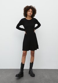 Schwarzes, geripptes Langarmkleid mit ausgestellt geschnittenem Saum, kombiniert mit klobigen schwarzen Stiefeletten, die ein strukturiertes Strickdesign betonen.