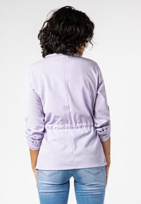 Světlý fialový top s dlouhým rukávem, zdobený řasením v pase a s volnějším střihem, doplněný o přiléhavé modré džíny, zobrazený ze zadní strany.