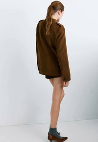 Veste en laine marron avec un col haut, des boutons en décoration et une coupe ample, associée à un short noir et des chaussures marron à talons. Chaussettes grises visibles.