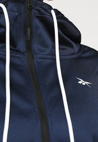Sweat à capuche zippé bleu marine avec cordons blancs et petit logo blanc Reebok sur la poitrine, vu de face.