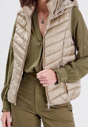 Femme portant une veste matelassée sans manches beige sur un chemisier vert olive et un pantalon, avec des colliers en or superposés et des bagues.