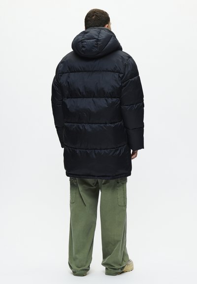 Man seen from behind wearing a black puffer jacket with hood, loose green cargo pants, and beige shoes standing against a white background.