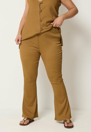 Femme portant un pantalon en maille côtelée moutarde et un top sans manches boutonné assorti, avec des sandales marron, debout devant un fond clair uni.