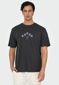 Mørkegrå t-shirt med korte ærmer, der har et "OSAKA" grafik i hvid og lilla, med en afslappet pasform og blødt bomuldsstof.