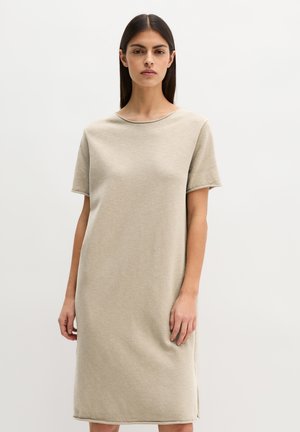 Vrouw met lang donker haar, gekleed in een eenvoudige, beige jurk met korte mouwen die tot op de knie valt, tegen een effen witte achtergrond.