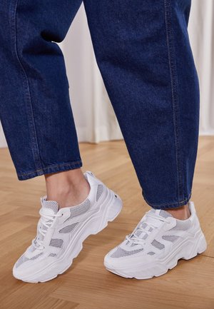 Persona con vaqueros azules y zapatillas blancas gruesas de pie sobre un suelo de madera clara en el interior.