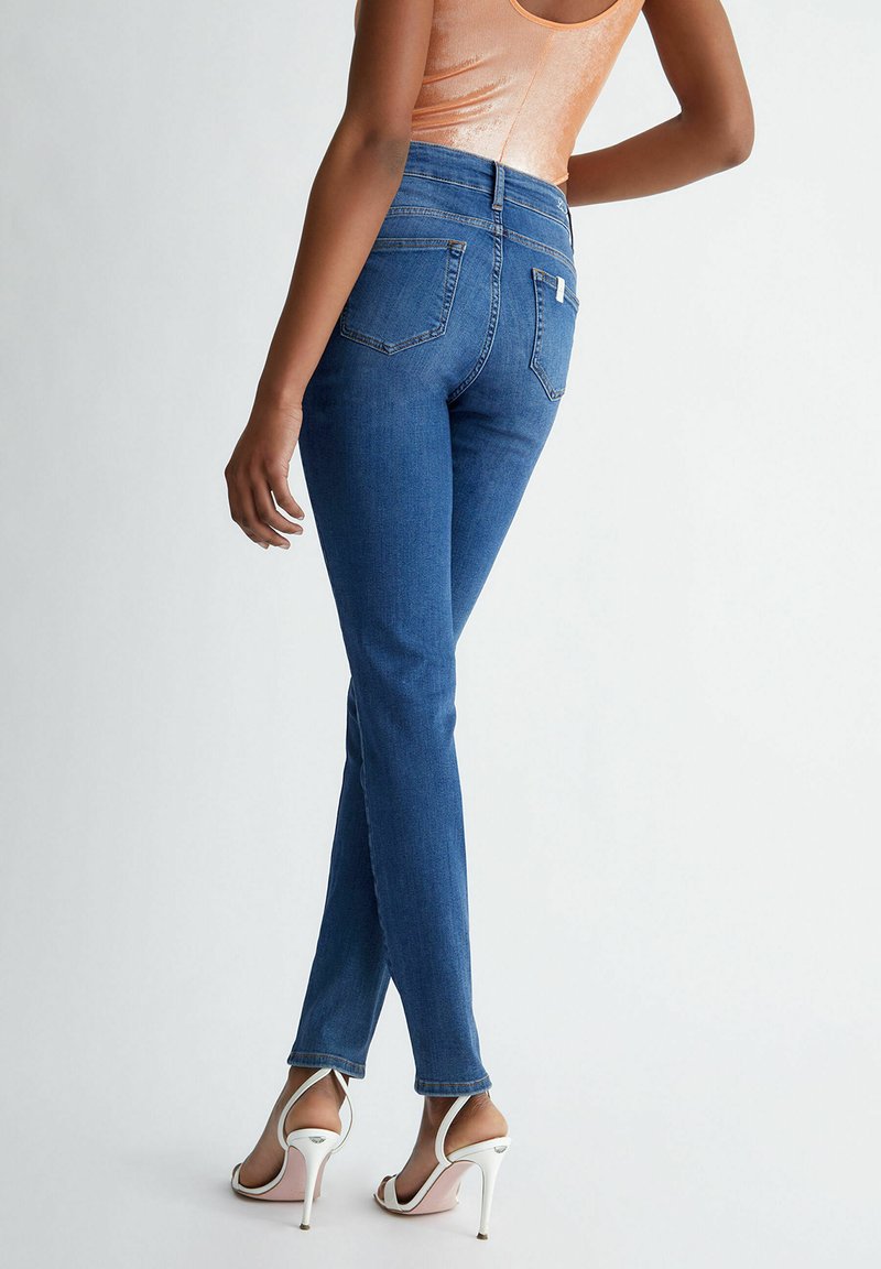 Jean bleu taille haute avec une coupe slim, dotés de poches arrière et de coutures proéminentes. Portés avec des talons aiguilles blancs à brides. Texture lisse.