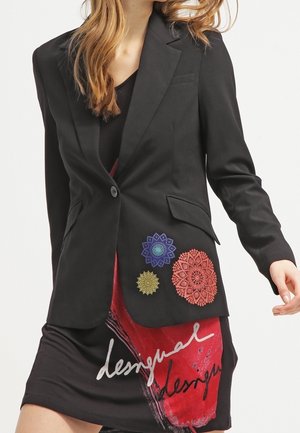 Nainen pukeutuneena mustaan bleiseriin, jossa on värikkäitä pyöreitä kirjailuja, yhdistettynä musta-punaiseen kuvioituun hameeseen, jossa on valkoista käsinkirjoitettua tekstiä.