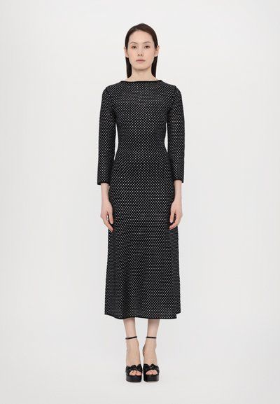 Abito nero a pois con maniche a tre quarti, corpino aderente e gonna fluida. Indossato con scarpe con plateau nere decorate con fiocchi.