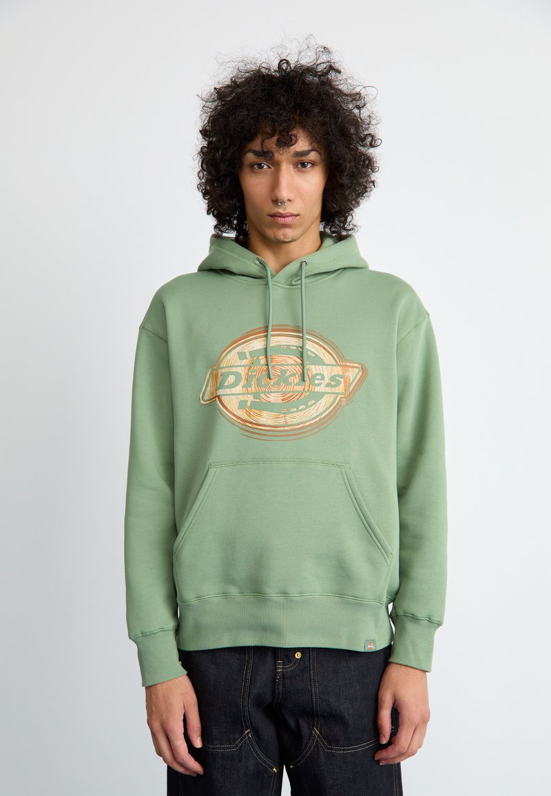 Hoodie de algodão verde com bolso frontal, grande impressão gráfica em laranja e bege, punhos e barra canelados. Usado com jeans escuros.