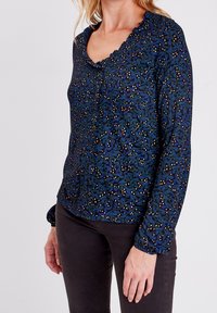 Dlouhá rukávová blůza s námořnickým pozadím, na kterém jsou zelené listové vzory a malé bílé tečky, nařasený límec a zapínání na knoflíky.