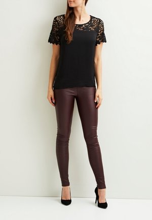 Kobieta w czarnej koronkowej bluzce z krótkim rękawem, spodniach w kolorze ciemnobrązowym o wyglądzie skóry oraz czarnych butach na wysokim obcasie stojąca na tle białej ściany.