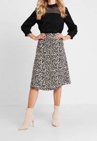 Schwarze Bluse mit strukturiertem Einsatz, kombiniert mit einem knielangen, beigen Leopardenmuster-Rock. Getragen mit beigen Knöchelstiefeln, vor einem schlichten Hintergrund.