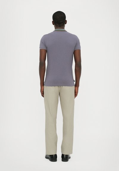 Camisa polo en púrpura claro con un cuello que presenta rayas verdes y blancas, combinada con pantalones beige claros y zapatos negros.