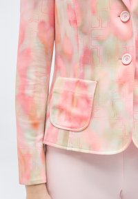 Παστέλ ροζ και ροδακινί σακάκι με σχέδια, κουμπιά και τσέπη με πατς στην αριστερή πλευρά, φορεμένο πάνω από ανοιχτό ροζ παντελόνια.