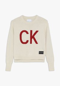Světle béžový pletený svetr s velkým červeným logem "CK" uprostřed a černou etiketou na dolní levé straně. Hladký výstřih a manžety.