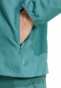 Chaqueta de color azul claro con puños elásticos, bolsillo lateral con cremallera y texto de marca sutil "AEROREADY" en la parte inferior frontal.