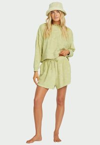 Mujer con cabello rubio largo, vistiendo una blusa de mangas largas texturizada en verde claro, pantalones cortos a juego y un sombrero de cubo, de pie descalza sobre un fondo liso.
