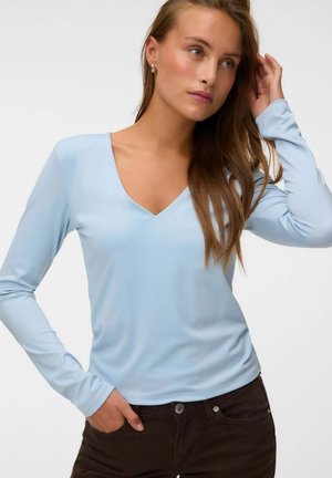 Femme aux longs cheveux bruns portant un haut bleu clair à col en V à manches longues et un pantalon foncé, posant avec une main dans la poche et l'autre touchant ses cheveux.