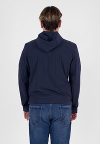 Giacca a vento blu navy con cappuccio realizzata in materiale liscio, con un fit rilassato e una superficie leggermente testurizzata, abbinata a jeans blu in denim.