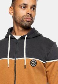 Sudadera con capucha y cremallera, con un diseño en bloques de color en tonos beige y gris oscuro. Presenta un parche de logo redondo, cordones blancos y una textura suave.