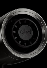 Gros plan sur un appareil circulaire noir avec une bordure métallique et le logo « ghd » au centre, sur un fond noir.