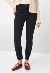 Marineblauwe, nauwsluitende broek gemaakt van rekbare stof, voorzien van een ritssluiting en knoop, zijzakken en een gladde textuur. Draagt bijpassende bordeauxrode schoenen.