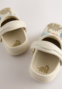 Sabots beige clair avec brides de talon ajustables, décorés de breloques colorées en forme de dinosaures et de nature sur le dessus et à l'intérieur des semelles.