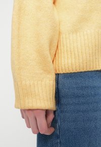 Main gauche reposant à côté du corps, portant un pull en tricot jaune pâle avec un poignet côtelé, assorti à un jean en denim bleu.