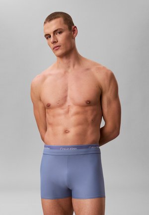 Man zonder shirt met kort haar, gekleed in blauwe Calvin Klein boxershorts, staand met zijn handen achter zijn rug tegen een effen grijze achtergrond.