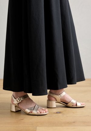 Gold strappy sandals with block heels. Features crisscross straps and ankle buckle. Worn with a long black skirt, on a wooden floor.