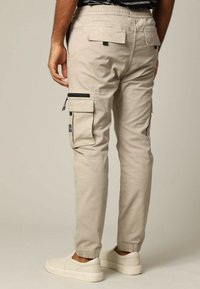 Beige Cargo-Hosen mit elastischem Bund, schmal zulaufendem Bein, seitlichen und hinteren Taschen sowie schwarzen Reißverschlussakzenten. Hergestellt aus einer Baumwollmischung.