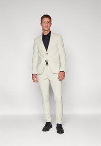 Világos bézs öltöny, slim fit szabású, egygombos zakóval, fekete gombokkal, fekete inggel alatta és fekete öltönycipővel. Sima textúra.