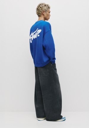 Blue sweatshirt with white graphic on the back, paired with wide-leg black jeans and blue-striped shoes. Smooth fabric, relaxed fit.