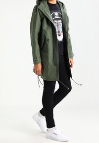 Parka à capuche vert olive avec cordons de serrage, t-shirt noir à graphisme avec un design de babouin, pantalon noir et baskets blanches.