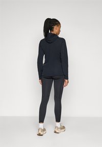 Marineblaue Hoodie mit taillierter Passform, kombiniert mit dunkelgrauen Leggings. Merkmale umfassen eine Kapuze, Nahtdetails und helle Schuhe.