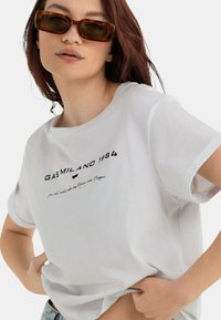 T-shirt bianca in cotone a maniche corte, con stampa nera "GAS MILANO 1984" e un fit casual, abbinata a occhiali da sole marroni.