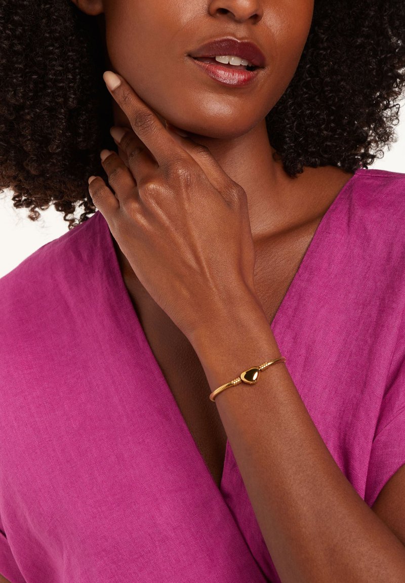 Gouden armband met een druppelontwerp en een gladde zwarte accent; gedragen op een hand met een subtiele lichtbruine nagellak tegen een roze achtergrond.