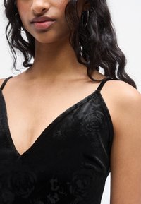 Φόρεμα από μαύρο βελούδο με V-λαιμόκοψη, λεπτές τιράντες και διακριτικό λουλουδένιο μοτίβο. Απαλή υφή με εφαρμοστή γραμμή.