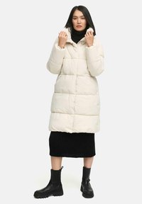 Light beige puffer coat with a high collar and zip closure. Features quilted texture, side pockets, and a relaxed fit. Worn over a black ribbed dress.