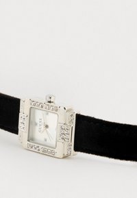 Relógio retangular prateado com mostrador branco, bracelete de camurça preta e realces de cristal transparente na luneta. Nome da marca "GUESS" exibido.
