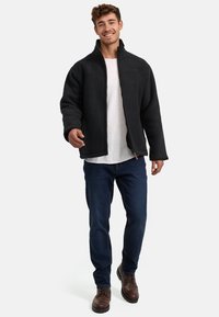 Chaqueta de forro polar negra con cuello alto y cierre de cremallera, combinada con una camiseta blanca, vaqueros azules y botas de cordones marrones.
